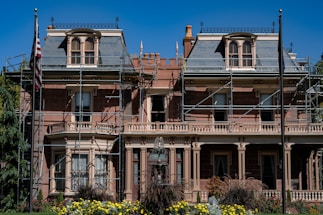 A large, historic-looking house with scaffolding around its structure, suggesting renovations or repairs. The building features ornate architectural details and is surrounded by vibrant greenery and colorful flowers. An American flag is visible on a flagpole in front of the house.