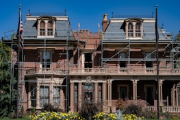 A large, historic-looking house with scaffolding around its structure, suggesting renovations or repairs. The building features ornate architectural details and is surrounded by vibrant greenery and colorful flowers. An American flag is visible on a flagpole in front of the house.