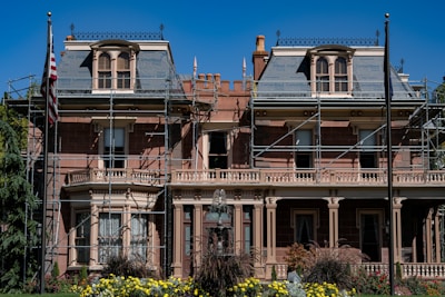A large, historic-looking house with scaffolding around its structure, suggesting renovations or repairs. The building features ornate architectural details and is surrounded by vibrant greenery and colorful flowers. An American flag is visible on a flagpole in front of the house.