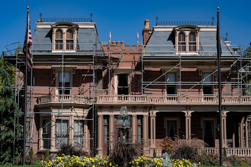 A large, historic-looking house with scaffolding around its structure, suggesting renovations or repairs. The building features ornate architectural details and is surrounded by vibrant greenery and colorful flowers. An American flag is visible on a flagpole in front of the house.