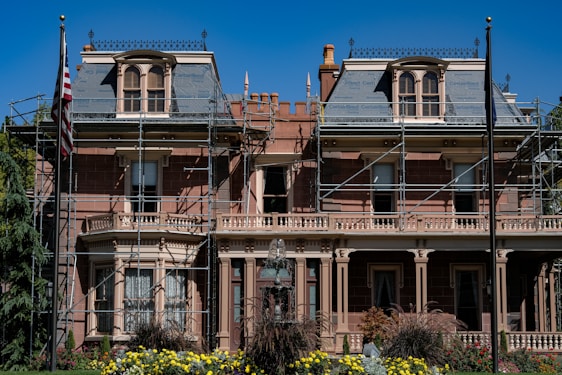 A large, historic-looking house with scaffolding around its structure, suggesting renovations or repairs. The building features ornate architectural details and is surrounded by vibrant greenery and colorful flowers. An American flag is visible on a flagpole in front of the house.