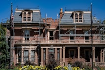 A large, historic-looking house with scaffolding around its structure, suggesting renovations or repairs. The building features ornate architectural details and is surrounded by vibrant greenery and colorful flowers. An American flag is visible on a flagpole in front of the house.