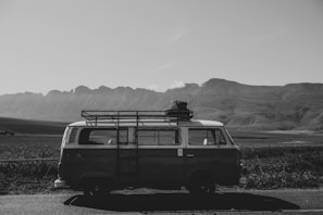 A vintage van is parked on a rural road with a scenic view of expansive fields and distant mountains in the background. The van has a rack on its roof carrying luggage, suggesting a road trip or adventure.