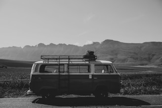 A vintage van is parked on a rural road with a scenic view of expansive fields and distant mountains in the background. The van has a rack on its roof carrying luggage, suggesting a road trip or adventure.