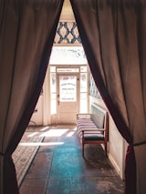 A vintage hotel entrance framed by curtains. The hallway features patterned wallpaper and a wooden bench with cushioned seating. Sunlight streams through the glass door, casting shadows on the tiled floor.