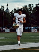 A football player wearing a white uniform with the number 5 on it is jogging on a field, holding a gold helmet in one hand. The sky is overcast with scattered clouds, and trees are visible in the background, along with a silhouetted building with a steeple.