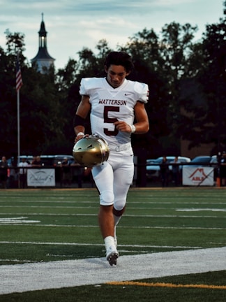 A football player wearing a white uniform with the number 5 on it is jogging on a field, holding a gold helmet in one hand. The sky is overcast with scattered clouds, and trees are visible in the background, along with a silhouetted building with a steeple.
