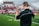 A coach wearing a headset stands on a football field, gesturing with his hand. He is dressed in a black Nike shirt, and bright green grass stretches out under a cloudy sky. In the background, there are blurred spectators sitting on the bleachers.