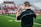 A coach wearing a headset stands on a football field, gesturing with his hand. He is dressed in a black Nike shirt, and bright green grass stretches out under a cloudy sky. In the background, there are blurred spectators sitting on the bleachers.