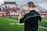 A coach wearing a headset stands on a football field, gesturing with his hand. He is dressed in a black Nike shirt, and bright green grass stretches out under a cloudy sky. In the background, there are blurred spectators sitting on the bleachers.
