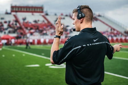 A coach wearing a headset stands on a football field, gesturing with his hand. He is dressed in a black Nike shirt, and bright green grass stretches out under a cloudy sky. In the background, there are blurred spectators sitting on the bleachers.