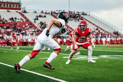 Two football players are on a green field, facing each other during a game. One player is in a white uniform with a black helmet, preparing to move, while the other is in a red uniform with a matching helmet, ready to defend. The stands in the background are filled with spectators, mostly wearing red.