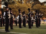 A marching band is performing outdoors on a grassy field. The band members are wearing black uniforms with plumed hats and are playing various brass and percussion instruments. The scene is set against a backdrop of trees and some spectators in the distance.