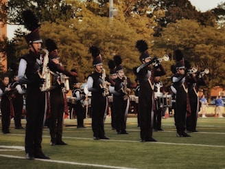 A marching band is performing outdoors on a grassy field. The band members are wearing black uniforms with plumed hats and are playing various brass and percussion instruments. The scene is set against a backdrop of trees and some spectators in the distance.