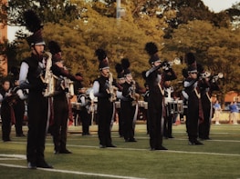 A marching band is performing outdoors on a grassy field. The band members are wearing black uniforms with plumed hats and are playing various brass and percussion instruments. The scene is set against a backdrop of trees and some spectators in the distance.