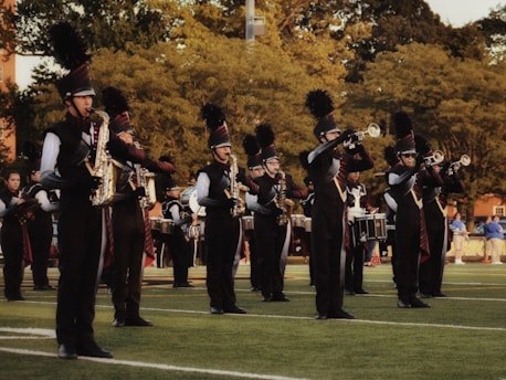 A marching band is performing outdoors on a grassy field. The band members are wearing black uniforms with plumed hats and are playing various brass and percussion instruments. The scene is set against a backdrop of trees and some spectators in the distance.