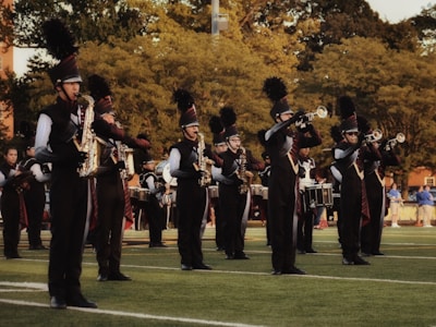A marching band is performing outdoors on a grassy field. The band members are wearing black uniforms with plumed hats and are playing various brass and percussion instruments. The scene is set against a backdrop of trees and some spectators in the distance.
