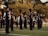 A marching band is performing outdoors on a grassy field. The band members are wearing black uniforms with plumed hats and are playing various brass and percussion instruments. The scene is set against a backdrop of trees and some spectators in the distance.