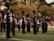 A marching band is performing outdoors on a grassy field. The band members are wearing black uniforms with plumed hats and are playing various brass and percussion instruments. The scene is set against a backdrop of trees and some spectators in the distance.