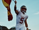 A football player wearing a white uniform with the number 5 and holding a flag stands against a clear blue sky, exuding confidence and readiness. The player wears a helmet, and the flag features yellow and dark red elements that complement the bold lettering on the uniform, which reads 'Watterson'. The setting hints at an outdoor field environment with trees visible in the background.