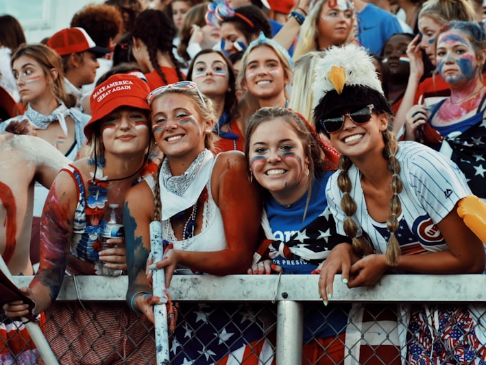 A group of young adults, mostly women, dressed in patriotic-themed clothing and face paint, standing behind a metal fence at what appears to be a sporting or public event. One wears a red hat with text and another is wearing an eagle hat. They are smiling and appear to be enjoying the event.