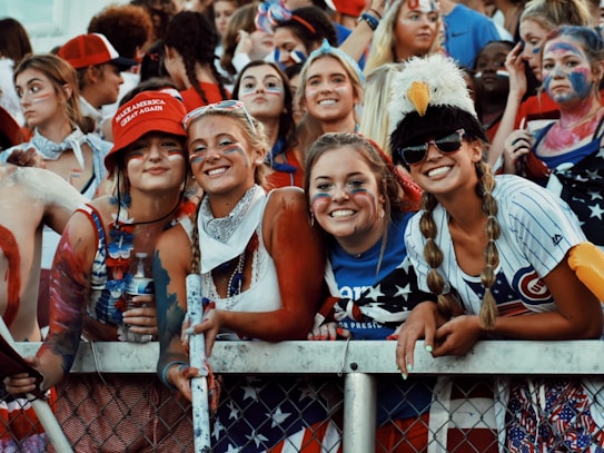 A group of young adults, mostly women, dressed in patriotic-themed clothing and face paint, standing behind a metal fence at what appears to be a sporting or public event. One wears a red hat with text and another is wearing an eagle hat. They are smiling and appear to be enjoying the event.