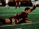 A soccer player in a maroon uniform is lying on a green turf field, appearing focused and determined. Other players in action are partially visible in the background in contrasting white uniforms.
