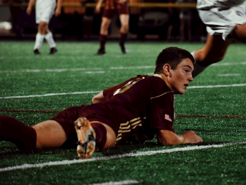 A soccer player in a maroon uniform is lying on a green turf field, appearing focused and determined. Other players in action are partially visible in the background in contrasting white uniforms.