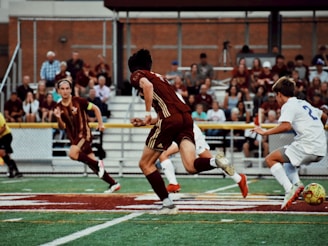 Players in soccer uniforms are actively engaged in a match on a green field with a maroon and white team playing against a team in white uniforms. Spectators are seated in the background on bleachers, watching the game unfold.