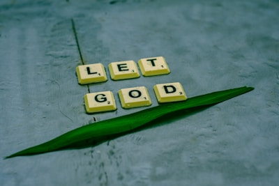 Scrabble tiles spelling out 'LET GOD' are arranged on a textured surface alongside a green leaf. The tiles are light yellow with black letters, providing a contrast against the muted background.