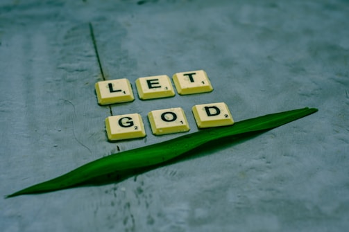Scrabble tiles spelling out 'LET GOD' are arranged on a textured surface alongside a green leaf. The tiles are light yellow with black letters, providing a contrast against the muted background.