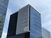 A modern, multi-story glass office building with a sleek, reflective facade. The structure features vertical white lines on one of its faces and has a large logo displayed at a high level. The sky in the background is clear and blue.