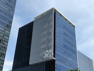 A modern, multi-story glass office building with a sleek, reflective facade. The structure features vertical white lines on one of its faces and has a large logo displayed at a high level. The sky in the background is clear and blue.