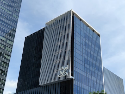 A modern, multi-story glass office building with a sleek, reflective facade. The structure features vertical white lines on one of its faces and has a large logo displayed at a high level. The sky in the background is clear and blue.