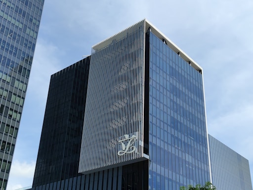 A modern, multi-story glass office building with a sleek, reflective facade. The structure features vertical white lines on one of its faces and has a large logo displayed at a high level. The sky in the background is clear and blue.