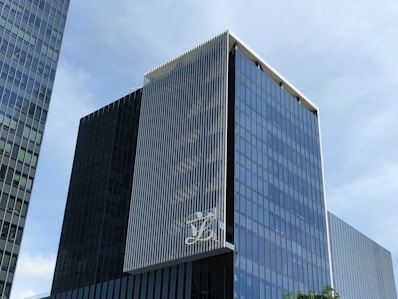 A modern, multi-story glass office building with a sleek, reflective facade. The structure features vertical white lines on one of its faces and has a large logo displayed at a high level. The sky in the background is clear and blue.