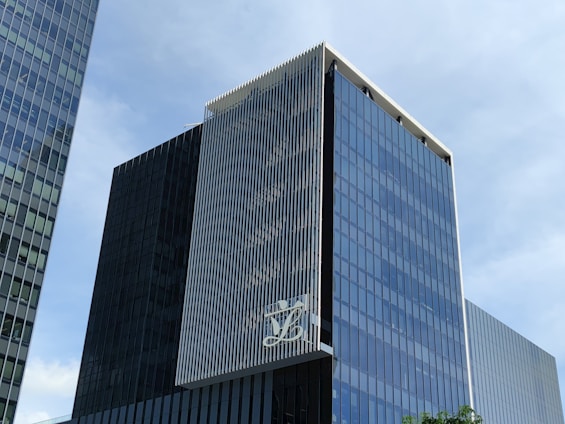 A modern, multi-story glass office building with a sleek, reflective facade. The structure features vertical white lines on one of its faces and has a large logo displayed at a high level. The sky in the background is clear and blue.