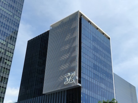 A modern, multi-story glass office building with a sleek, reflective facade. The structure features vertical white lines on one of its faces and has a large logo displayed at a high level. The sky in the background is clear and blue.