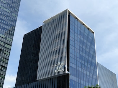 A modern, multi-story glass office building with a sleek, reflective facade. The structure features vertical white lines on one of its faces and has a large logo displayed at a high level. The sky in the background is clear and blue.