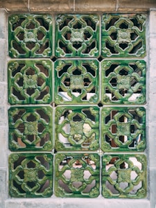 A wall composed of nine intricately designed green ceramic tiles, each featuring a floral pattern. The tiles are arranged in a three by three grid and are separated by thin seams of grout. The background behind the tiles is visible through the cut-out patterns, creating a sense of depth.