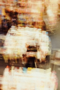 A blurred and abstract view of what appears to be a colorful indoor scene, possibly a convenience store or a market with various products on shelves. The image is out of focus, giving it a dreamlike or chaotic quality.