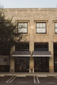 A commercial building with a weathered façade featuring large, grid-like windows. Black-and-white striped awnings cover the shopfronts below. The storefronts display clothing items behind the glass, and there are no people present. A row of outdoor string lights is strung across the building. The building is surrounded by trees, and a parking space is visible in the foreground.