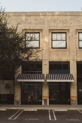 A commercial building with a weathered façade featuring large, grid-like windows. Black-and-white striped awnings cover the shopfronts below. The storefronts display clothing items behind the glass, and there are no people present. A row of outdoor string lights is strung across the building. The building is surrounded by trees, and a parking space is visible in the foreground.