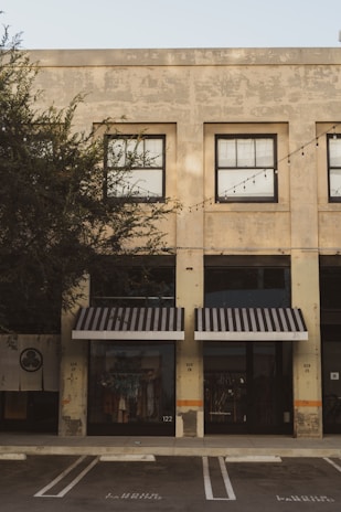 A commercial building with a weathered façade featuring large, grid-like windows. Black-and-white striped awnings cover the shopfronts below. The storefronts display clothing items behind the glass, and there are no people present. A row of outdoor string lights is strung across the building. The building is surrounded by trees, and a parking space is visible in the foreground.