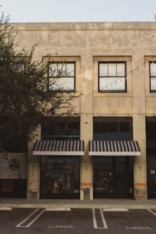 A commercial building with a weathered façade featuring large, grid-like windows. Black-and-white striped awnings cover the shopfronts below. The storefronts display clothing items behind the glass, and there are no people present. A row of outdoor string lights is strung across the building. The building is surrounded by trees, and a parking space is visible in the foreground.