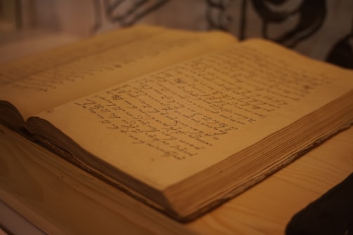 An antique book lies open, displaying handwritten text in a language that appears historical or script-like. The pages are yellowed with age, suggesting the book is old. The thick pages and binding add to the vintage feel, resting on a wooden surface.