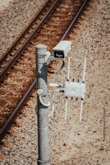 A surveillance camera is mounted on a metal pole beside railway tracks. The pole also has attached electronic equipment with antennas, likely for communication or data transmission. The tracks run parallel on gravel-covered ground.
