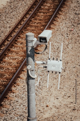 A surveillance camera is mounted on a metal pole beside railway tracks. The pole also has attached electronic equipment with antennas, likely for communication or data transmission. The tracks run parallel on gravel-covered ground.