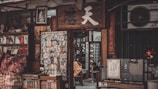 A small, cluttered shop entrance with various traditional items displayed. Books and papers are stacked on shelves, and a cloth bag with red patterns lies in front. A wooden sign with Chinese characters hangs above the entrance, and an assortment of posters and images are visible. Inside, a person is seated, possibly reading or using a device.