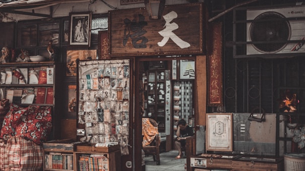 A small, cluttered shop entrance with various traditional items displayed. Books and papers are stacked on shelves, and a cloth bag with red patterns lies in front. A wooden sign with Chinese characters hangs above the entrance, and an assortment of posters and images are visible. Inside, a person is seated, possibly reading or using a device.